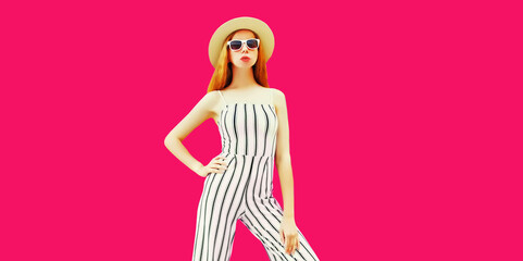 Stylish model woman wearing summer round straw hat, white striped jumpsuit posing on pink background