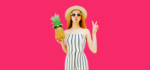 Summer portrait of beautiful young woman with pineapple blowing her red lips sending sweet air kiss wearing straw hat, striped jumpsuit on pink background