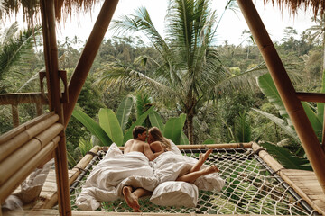 Young caucasian travel couple lie  in hammock in  bamboo eco house, nature and mountains on background. Morning in tropical resort on Bai island