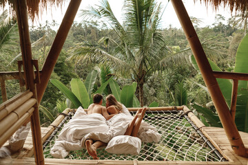 Happy travel couple on hammock balcony of bamboo tree house with jungle nature view. Vacation in beaitiful hidden place, honeymoon on Bali island - obrazy, fototapety, plakaty