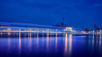 Fototapeta na wymiar Moored Containervessel at Terminal Burchardkai in City of Hamburg during night time with blue colored light stripes from a tourism boat on the river Elbe. Long exposure shot.