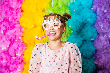 Cute girl celebrating birthday, wearing party paper glasses, blowing party horns, laughing and...
