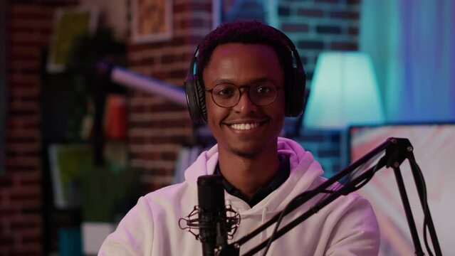 Portrait of african american podcaster smiling confident at camera while recording podcast in home studio. Male online radio host posing happy while talking live to audience using microphone.