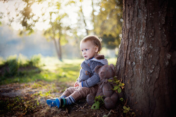 Malutki chłopiec siedzi pod drzewem w parku razem ze swoim misiem i patrzy na wodę