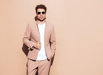 Portrait of handsome confident stylish hipster lambersexual model. Sexy modern man dressed in elegant beige suit. Fashion male with curly hairstyle posing in studio. Isolated. In sunglasses