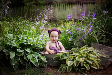 Malutki chłopiec w czapeczce misia siedzi w ogrodzie pomiędzy fioletowymi kwiatami i pięknie się uśmiecha