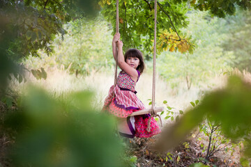 Dziewczynka w letniej sukience huśta się na huśtawce powieszonej na drzewie, śmieje się i...