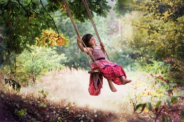 Fototapeta Dziewczynka w letniej sukience huśta się na huśtawce powieszonej na drzewie, śmieje się i śpiewa piosenki wymachując nogami obraz