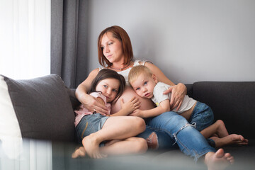 Kobieta w ciąży ze swoimi dziećmi siedzi w domu na kanapie, mama przytula w objęciach swoje dzieci - obrazy, fototapety, plakaty