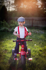 Mały chłopiec uczy się jeździć na rowerku w pięknej retro stylizacji i czerwonych szelkach, ogród w wakacyjnych promieniach słonca
