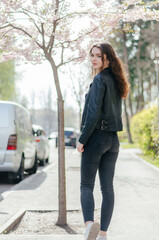 girl in black jeans and a leather jacket walks on the sidewalk near the sakura tree