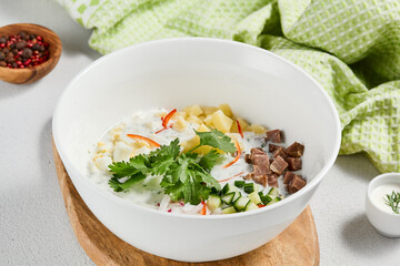 Traditional russian soups - okroshka with fresh vegetables, meat and kefir. Cold summer soup on white table. Aesthetic composition with traditional russian food and spices, textile