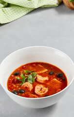 Fish tomato soup with shrimps and black olives. Seafood soup with prawns in white ceramic bowl on concrete background. Fish dish with seafood. Tomato soup in minimal composition.
