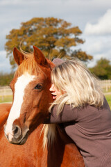 Close up shot of a pretty,  long haired blond girl hugging her chestnut pony outdoors in its field.