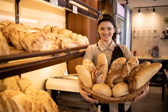 Portrait of beautiful female deli worker in uniform holding fresh pastries and bred in supermarket bakery department.