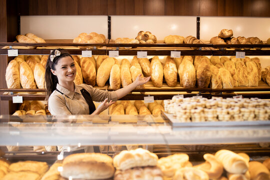 Portrait of female bakery worker showing fresh baked pastries and bread on shelf in bakery shop.