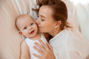 Fototapeta na wymiar Happy young mother with baby at home in white room, mom and baby are lying, kissing the baby at home on the bed in the bedroom