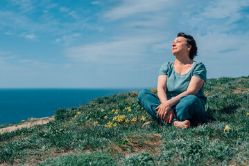 Naklejka na ściany i meble an adult woman, pensioner sits on the grass, legs crossed in a lotus position barefoot with a smile on her face on a sunny day on a mountain against the backdrop of the sea in summer, spring
