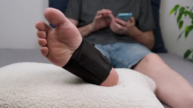 man after an ankle injury is lying on the couch with his smartphone. close-up of the injured leg in special fixing bandage in the ankle joint area. rehabilitation after injury. close-up. slow motion.