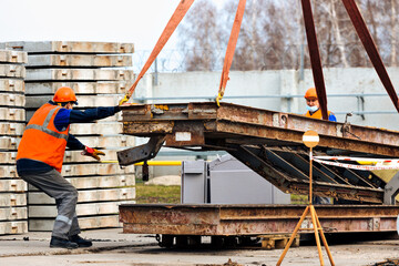 Naklejka premium Slinger in helmet and vest controls unloading of metal structures on construction site. White handyman unloads load.