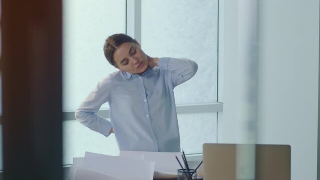 Woman Standing By the Table Massaging Back Suffering From Pain or Inflammation Processes Due to Long Sedentary Work at Office. Suffering Office worker Feeling Sick Because of all Work, Stress Backache