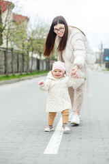Naklejka premium Little baby girl learns to walk with mom's help outside. Young mother and daughter toddler