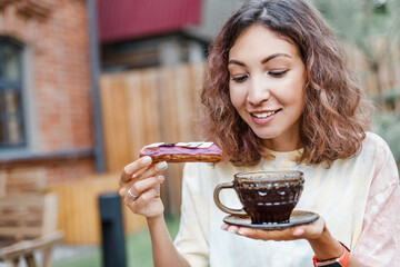 Happy woman eating delicious tasty eclair cake and drinking tea in outdoor cafe - obrazy, fototapety, plakaty