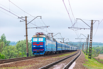 Fototapeta premium A powerful dual-system electric locomotive pulls a long intercity passenger train to the railway station. Spring cloudy weather.