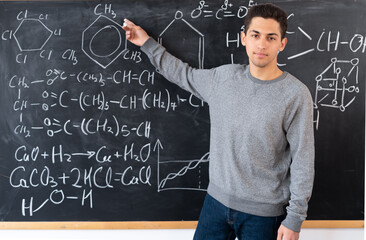 the teacher writes on the blackboard various formulas for chemistry in high school. a young Arab teacher points his finger at the formulas. - Powered by Adobe