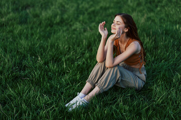 A beautiful girl relaxing in nature sitting in pants and top on the green grass in the rays of the setting summer sun. The concept of health and care for the body and the environment 