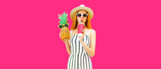 Summer portrait of beautiful woman drinking juice and holding pineapple wearing straw hat, striped jumpsuit, sunglasses on pink background