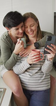 Vertical video of happy caucasian lesbian couple drinking coffee and using smartphone at home