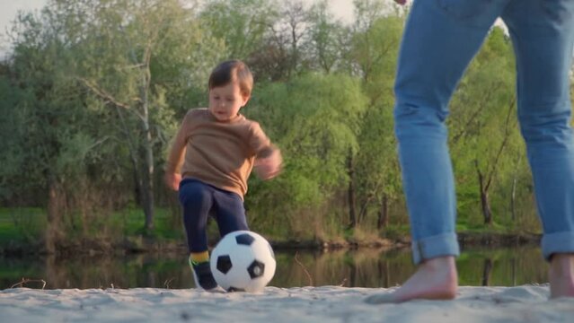 Little Kid Kicks Soccer Ball to His Father. Slow Motion. Young Man And Little Boy Playing With Black and White Football Ball Outdoors