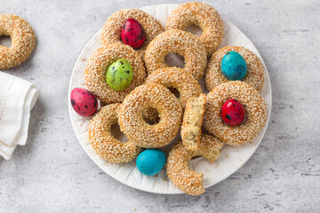 White plate with traditional Greek Easter cookies Koulourakya or Koulourya shortbread rings with sesame seeds, decorated with colored eggs on a gray textured background. Homemade Easter pastries