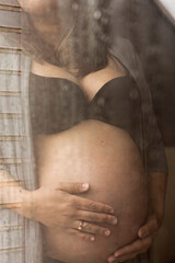 Nine months pregnant woman caresses her belly with her hands looking out the window at home on a rainy day thinking about her baby. The mother wears her ring and black bra, Concept of pregnancy. 