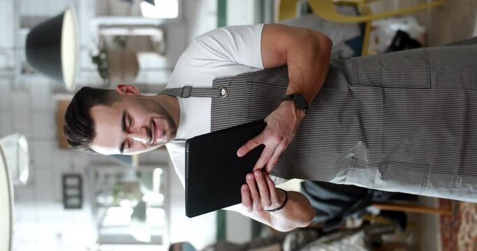 Vertical footage of handsome Caucasian young man waiter in apron standing in cafe kitchen and using tablet device. Male chef of restaurant texting, tapping and scrolling on computer screen.