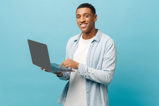Optimistic african-american male student in casual jeans shirt using laptop pc isolated on blue background, smiling multiracial freelancer man looking enjoying remote work, advertising concept