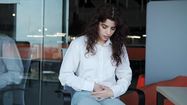 Beautiful freelance girl in a white shirt with curly hair sits on a chair in the office and suffers from abdominal pain. A woman with menstrual pain or poisoning, suffers from abdominal cramps.