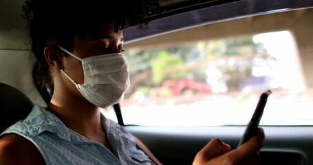 Passenger girl in backseat taxi wearing mask looking at smartphone