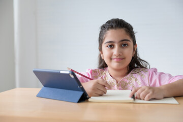 portrait cute Indian girl writing on notebook and online learning class from tablet, education concept