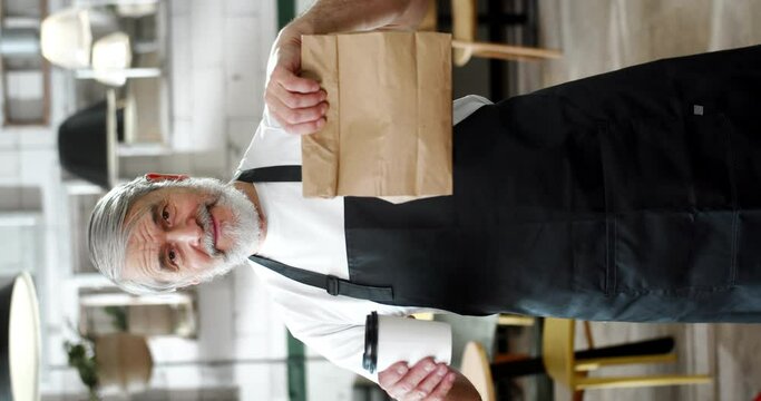 Vertical portrait of Caucasian old man waiter in apron handing coffee and package to-go to camera. Senior grey-haired male worker of coffee shop giving drink and food purchase to client. Vertically.