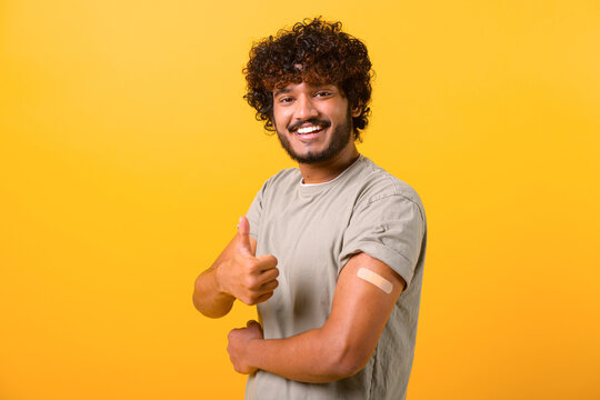 Smiling young curly Indian guy showing arm with band-aid after vaccine injection isolated on yellow background, vaccinated latino man showing thumb up