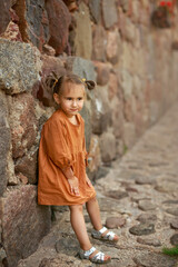 A cute girl with funny tails in a dress is standing against the wall of a masonry building. antique stone building. A thoughtful child looks away with a smile