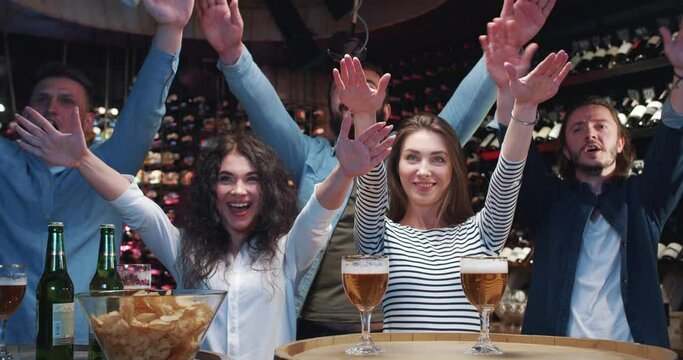 Caucasian sport fans doing cheerleading dance moves in bar while watching game. Men and women friends supporting sport team and cheering up in evening in front of TV in pub.