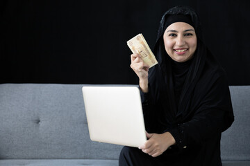 portrait muslim woman holding cash money and laptop computer on black background