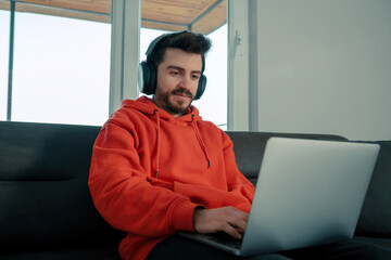 Young man wearing red hoodie sitting on couch in living room at home enjoying studying using laptop and headset looking at device screen listening audio making some notes. Working from home. - Powered by Adobe