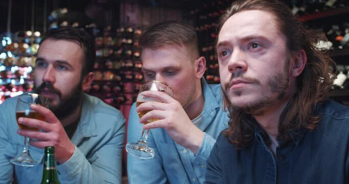 Caucasian handsome guys drinking beer in pub at night, talking and watching sport match. Mest males friends sipping drinks and cheering up for game on sport channel. Footbal fans in bar.