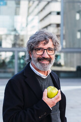 Elegant mature businessman with glasses eating apple outside the office building.
