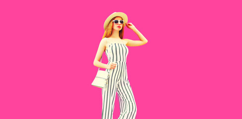 Stylish model woman wearing summer round straw hat, white striped jumpsuit posing on pink background