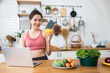 Smiling strong fitness woman wearing sport suit with laptop and chili pepper, vegetables in the kitchen. Healthy diet and sport concept. Finding information for diet